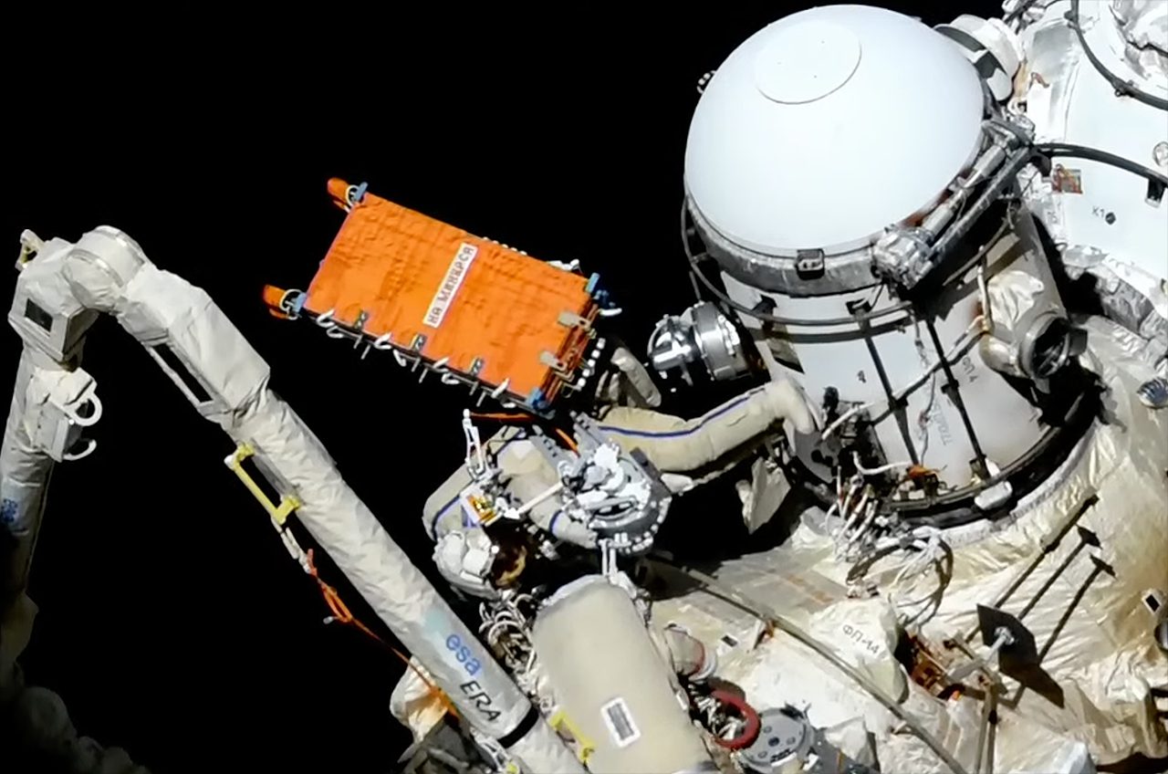 Un astronauta vestido con un traje espacial blanco instala un sistema de comunicaciones por radar naranja en la Estación Espacial Internacional.