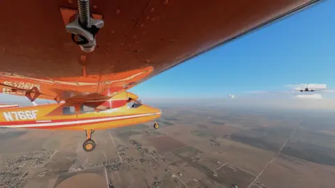 Un pequeño avión naranja vuela, siendo remolcado por otro avión delante de él.