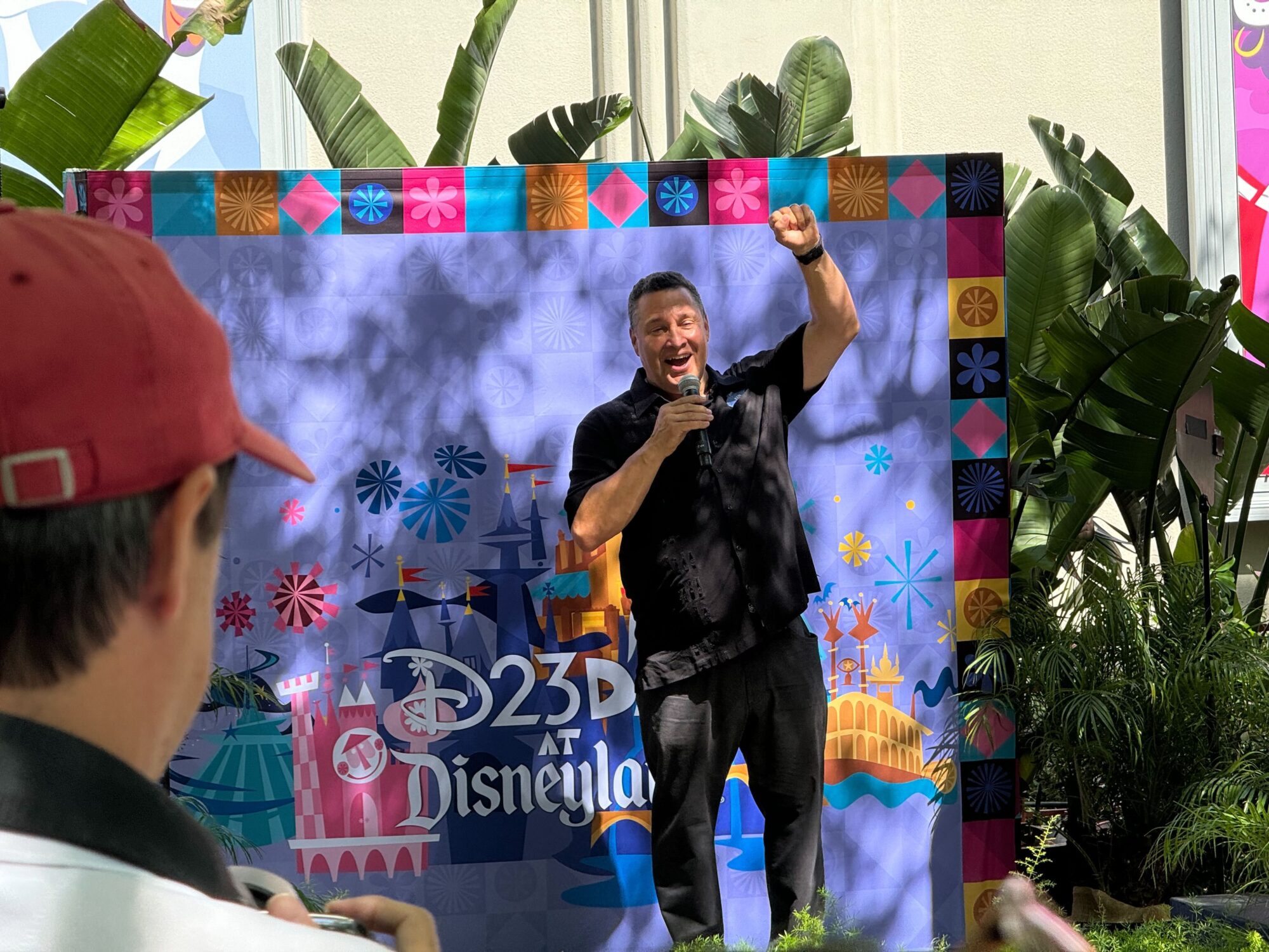 Hombre parado frente a un fondo colorido con "D23 en Disneylandia" Text, sosteniendo un micrófono y levantando el puño derecho, habla ante una pequeña audiencia en un evento al aire libre.