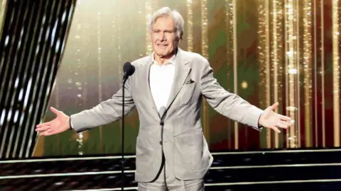 Reuters Harrison Ford saluda mientras es homenajeado en los Disney Legends Awards 2024 durante la Disney's D23 Expo en Anaheim. Harrison es un hombre de 82 años con cabello gris. Con un traje gris pálido sobre una camisa blanca, se encuentra en el escenario con los brazos extendidos.