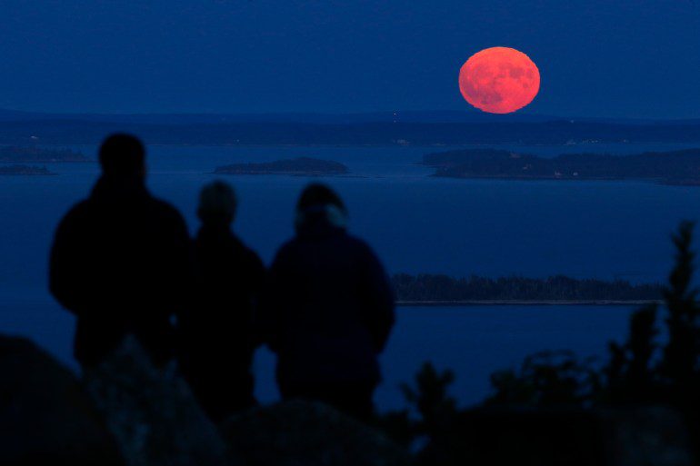 Tres personas observan aparecer la luna de cosecha casi llena sobre la bahía de Penobscot