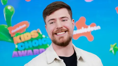 Imágenes de Jimmy Donaldson, también conocido como MrBeast, en los Nickelodeon Kids' Choice Awards. MrBeast tiene cabello castaño corto y barba corta. Viste una chaqueta blanca sobre una camisa negra y se para frente a un letrero azul brillante con la marca naranja de Nickelodeon. 