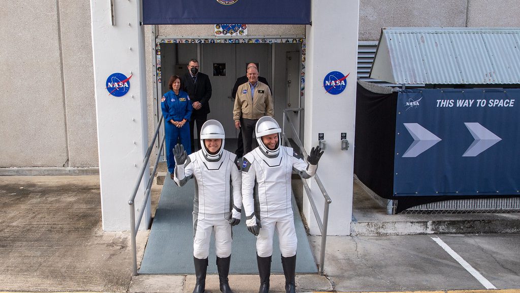 Los astronautas de la tripulación de nueve personas con trajes espaciales blancos y negros saludan mientras se dirigen a la plataforma de lanzamiento.