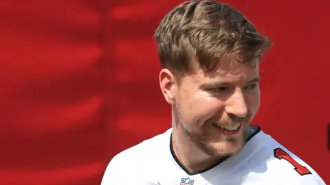 Jimmy Donaldson, conocido como Mr. Best, tiene cabello castaño corto y barba corta. Viste un uniforme blanco de fútbol americano con un único número rojo en el hombro izquierdo. Mira por encima de su hombro izquierdo lejos de la cámara y aparece en la foto frente a un fondo rojo. 