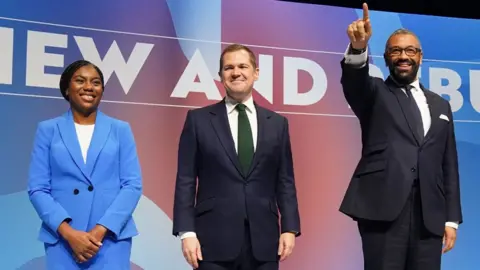 PA Media Los dos candidatos restantes, Kemi Badenoch con un traje azul brillante, Robert Jenrick con un traje azul marino y Cleverly con un traje azul marino, estaban uno al lado del otro en el escenario de la conferencia del Partido Conservador, todos sonriendo al público.